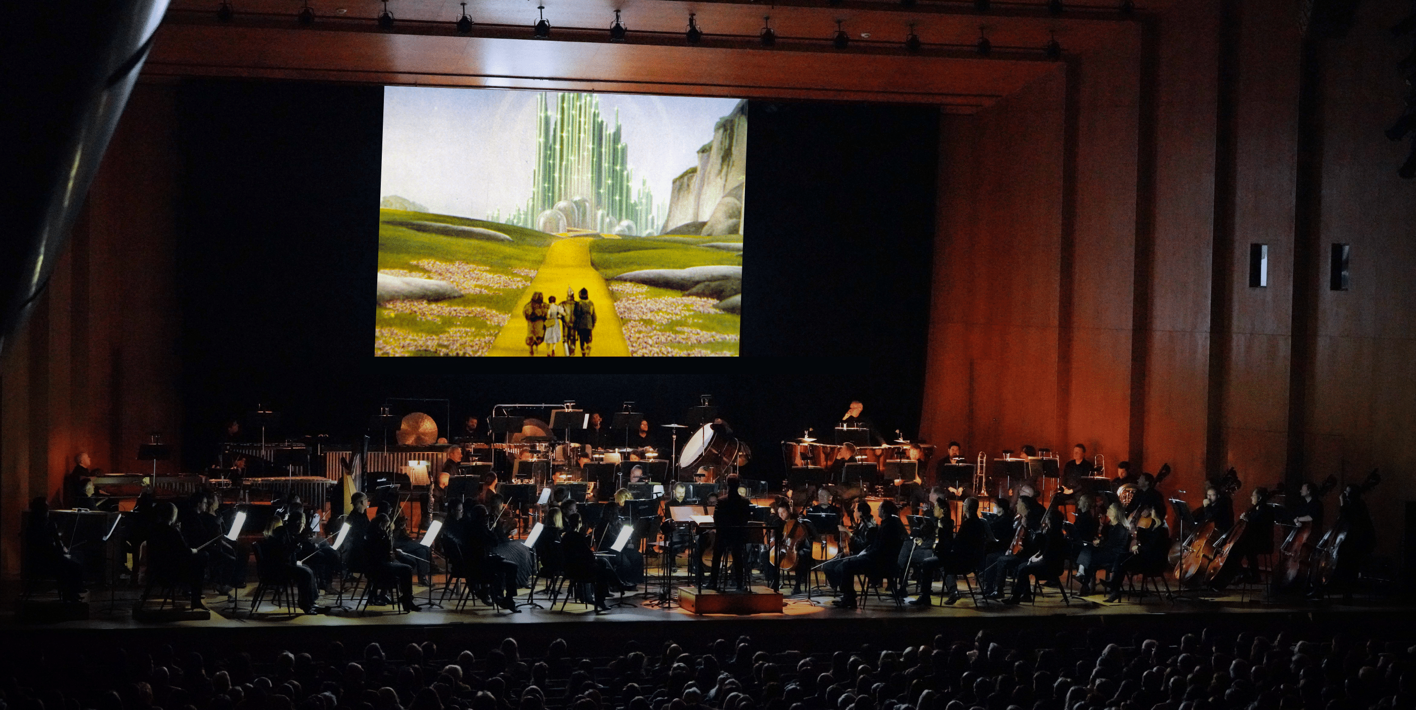 A symphony orchestra performs on stage beneath a large screen showing a scene from The Wizard of Oz with Dorothy and friends walking toward the Emerald City on the yellow brick road.