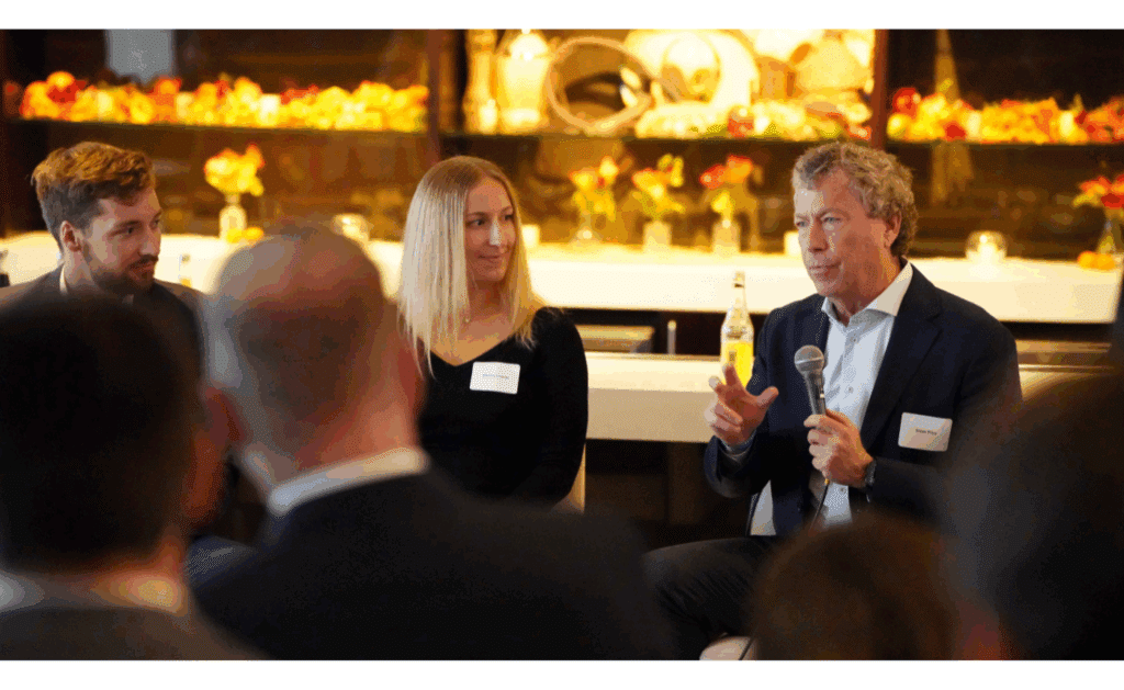 Three people sit and converse at an event. The man on the right, holding a microphone, speaks while the other two listen. Warm lighting and flower arrangements decorate the background. Audience members’ heads are visible in the foreground.