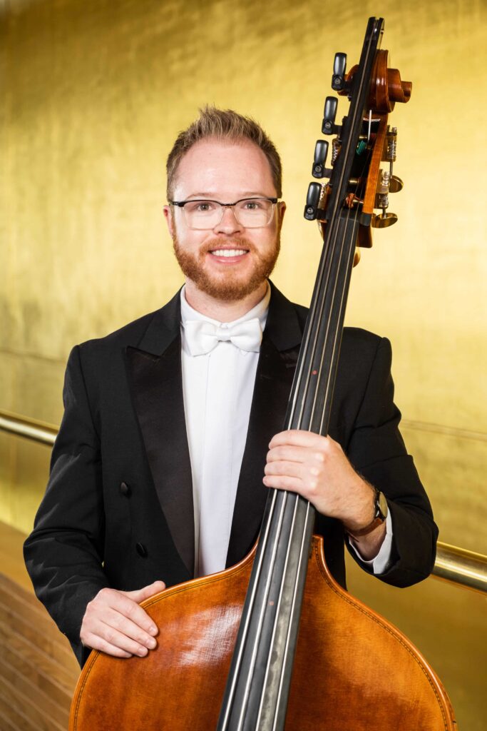 A man in formal attire with a white bow tie and glasses stands smiling, holding an upright bass against a golden background.