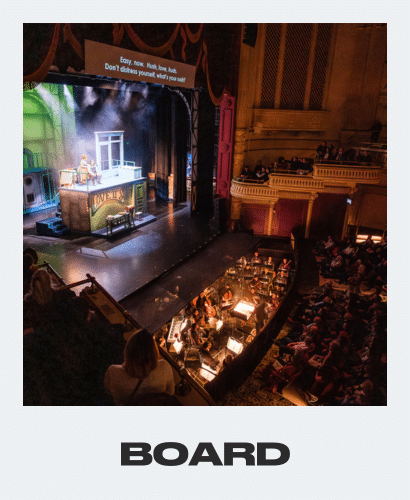 A musical theater performance on stage, with actors and set pieces under bright lights. An orchestra plays in the pit below, while audience members watch from theater seats and balconies. The word BOARD appears at the bottom.