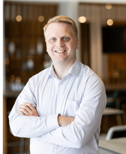 A man with short blond hair wearing a light-colored, long-sleeve button-up shirt stands indoors with his arms crossed, smiling at the camera. The background is softly blurred, showing a modern office setting.
