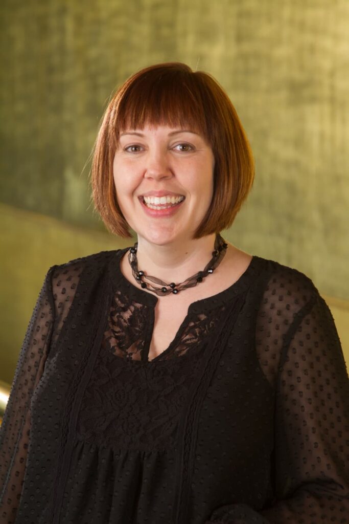 A woman with short auburn hair and bangs, wearing a black sheer blouse and beaded necklace, smiles in front of a blurred yellow-green background.