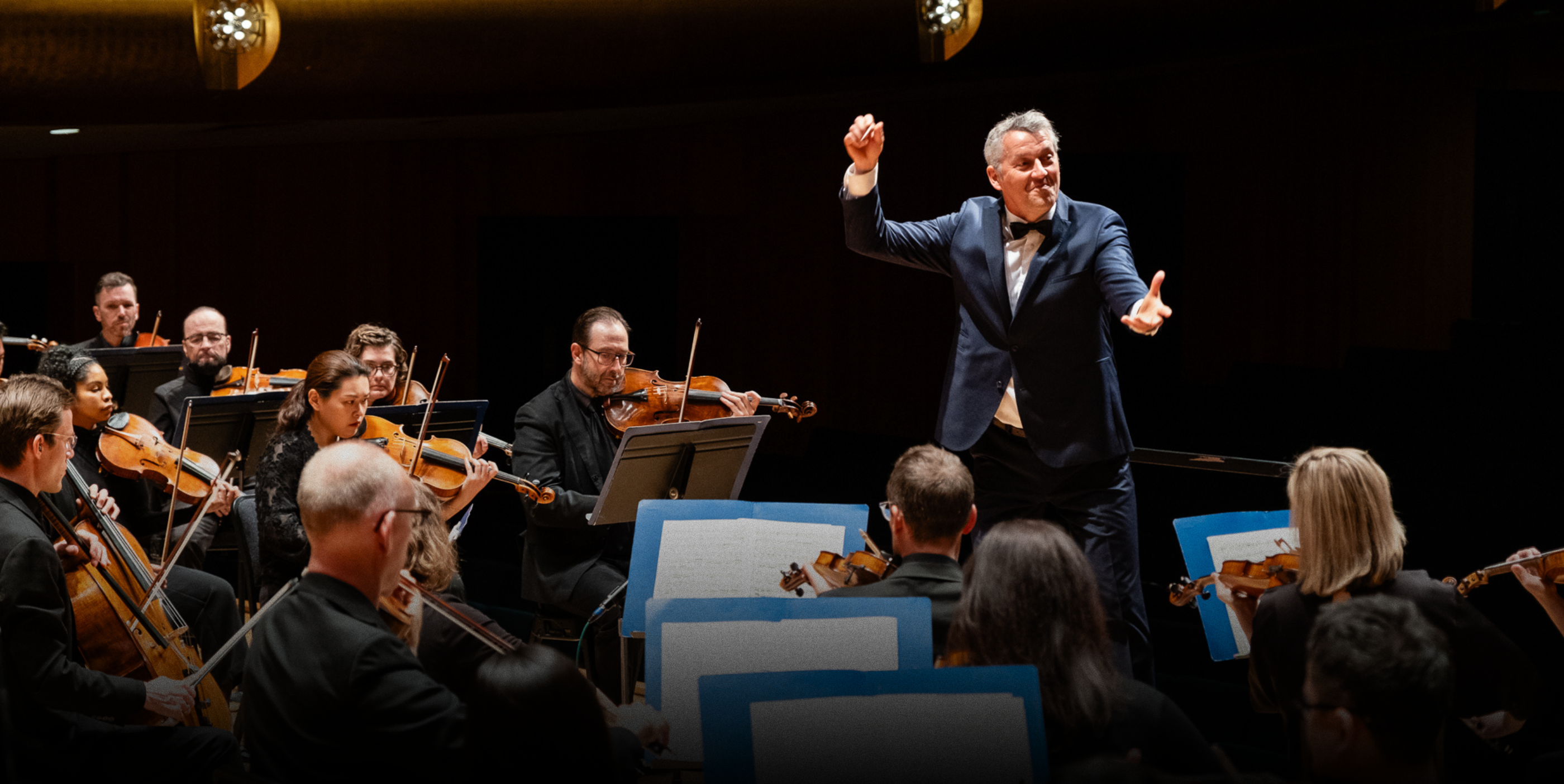 A conductor enthusiastically leads an orchestra of musicians playing string instruments on stage during a concert, with music stands and sheet music visible.
