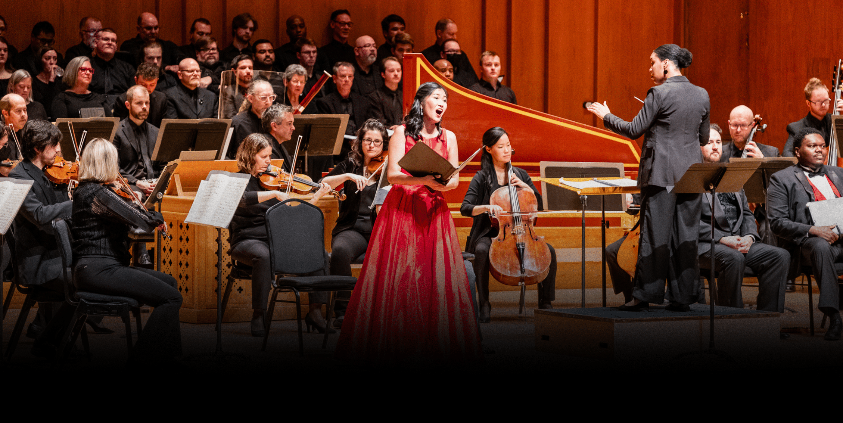A female soloist in a red dress sings passionately on stage with a conductor and a seated orchestra behind her, featuring string and harpsichord musicians in a wood-paneled concert hall.