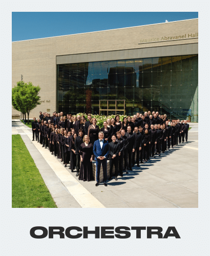 A large orchestra, dressed in black, stands in rows outside a modern building labeled Maurice Abravanel Hall. Lush green grass and trees surround the group, with a clear sky reflected in the building’s glass windows.