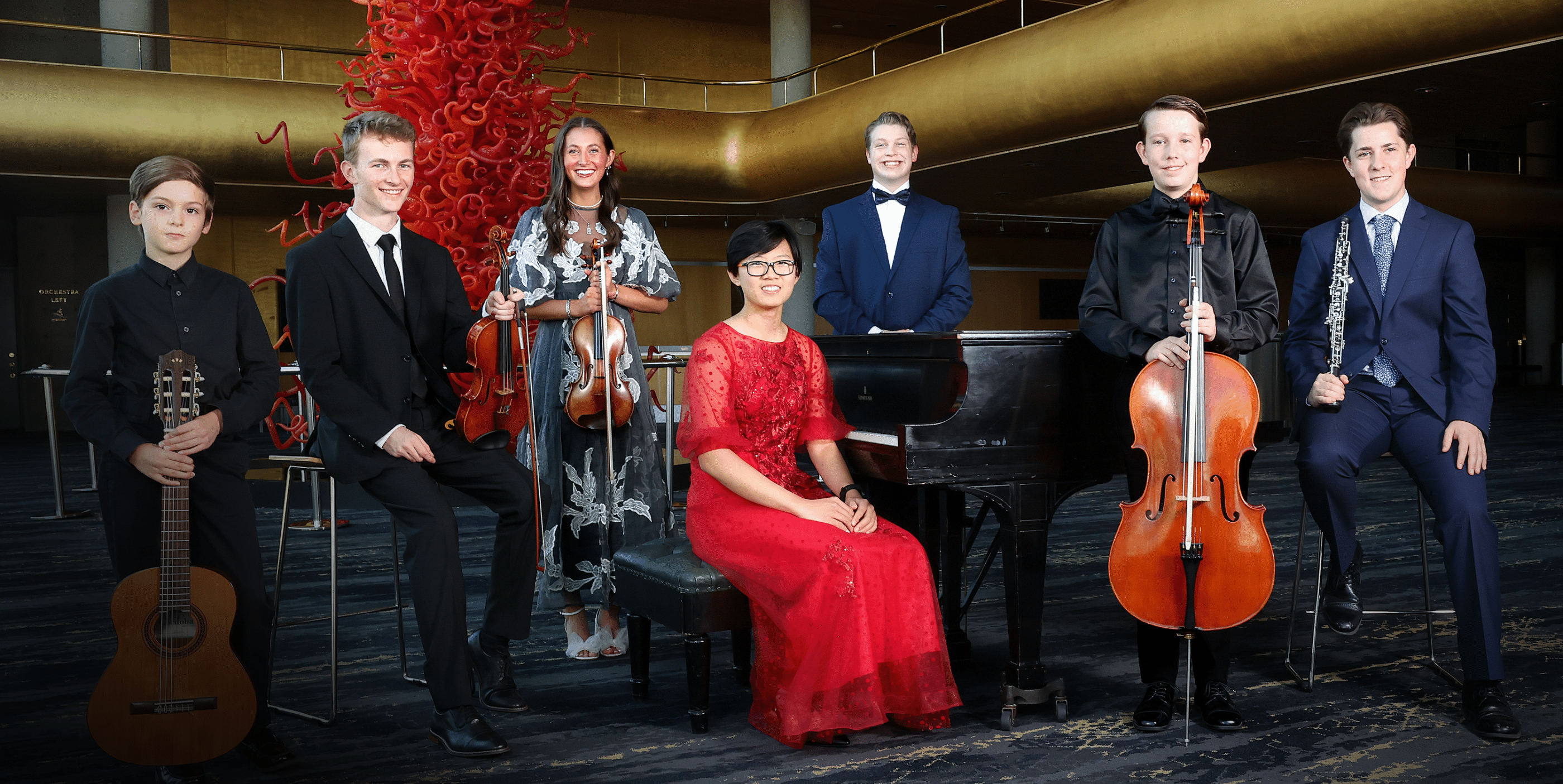 Seven young musicians in formal attire pose indoors with their instruments: guitar, violin, piano, cello, and clarinet. The pianist sits in a red dress at a grand piano; the others stand or sit nearby, smiling, with a red sculpture in the background.