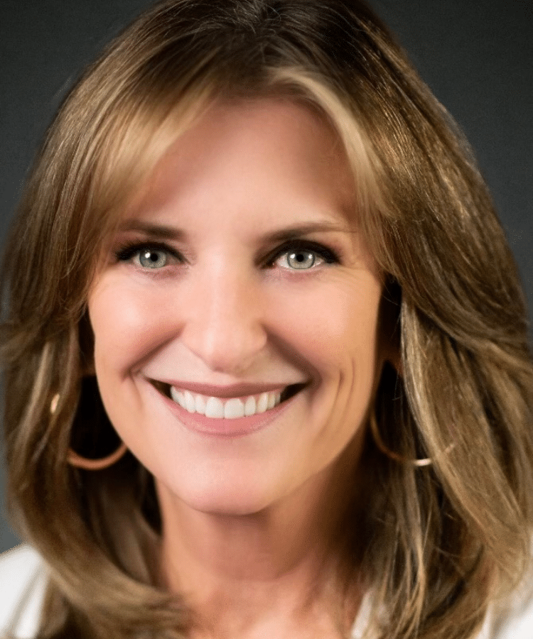 A woman with shoulder-length light brown hair, wearing hoop earrings and a white top, smiles warmly at the camera against a dark background.