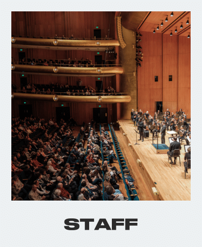A symphony orchestra performs on stage in a large concert hall with a seated audience and multiple balcony levels. The word “STAFF” is written in bold letters below the image.