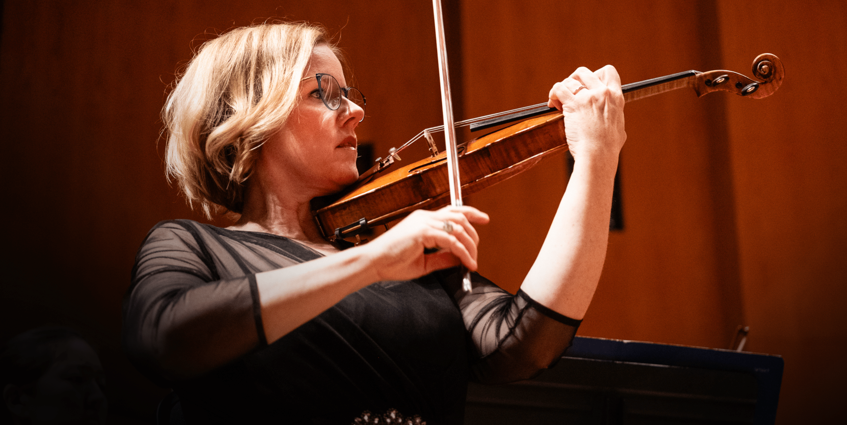 A woman wearing glasses and a black dress plays the violin on stage, focused on the music, with a warm brown background behind her.