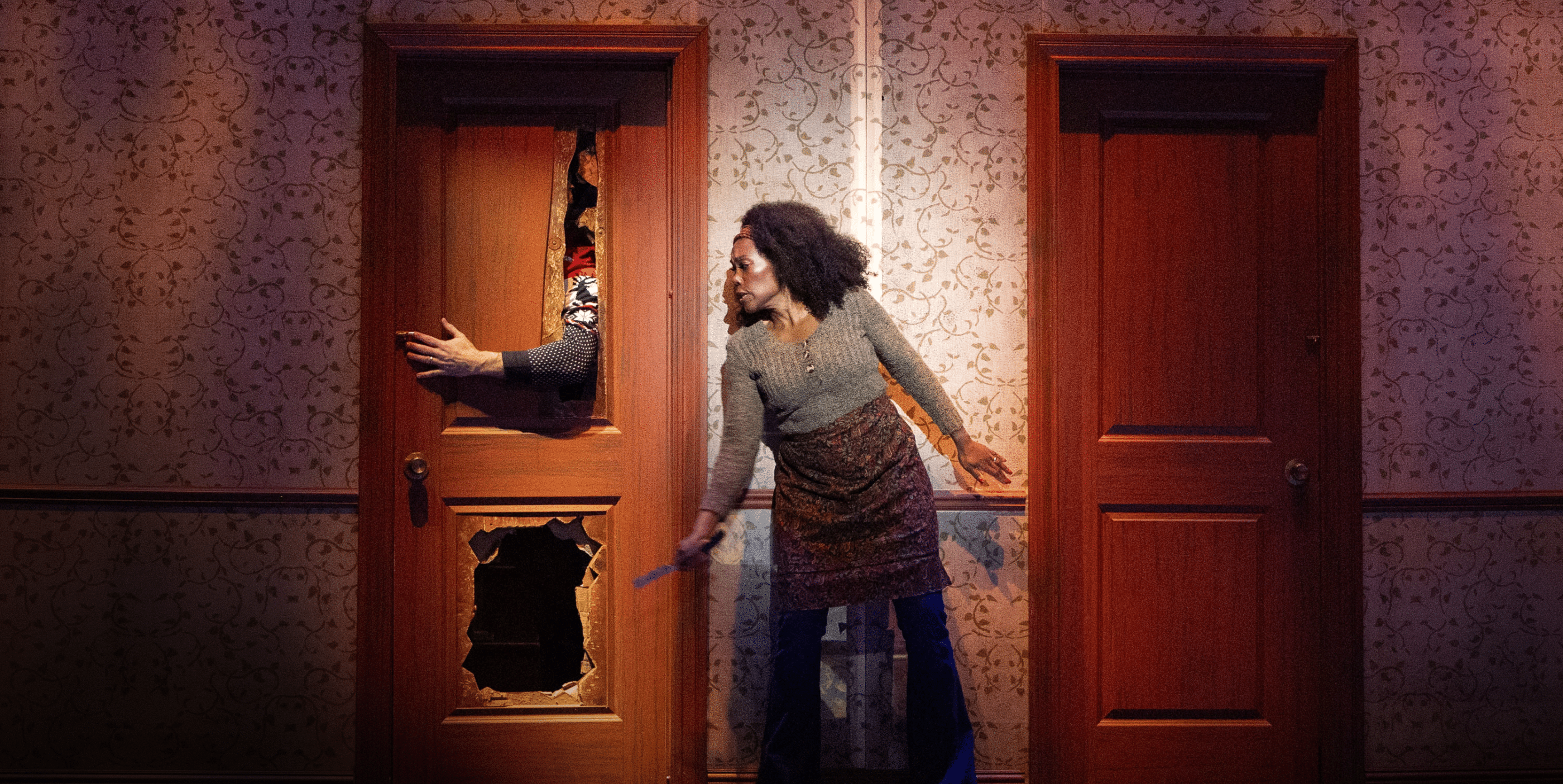 A woman cautiously opens a door while another person’s arm reaches through a broken panel in a nearby door, next to another closed door, in a dimly lit hallway with patterned wallpaper.