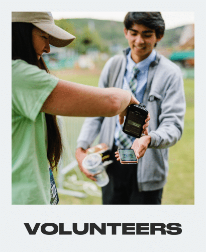 A volunteer scans a QR code on a young man’s phone outdoors. Both are smiling, and the man holds drinks and snacks. The word VOLUNTEERS is written in bold letters at the bottom.