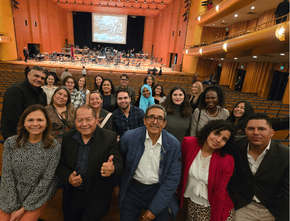 A group of people pose and smile in the foreground of a concert hall with a stage set for The Wizard of Oz performance. The hall has wooden walls and rows of empty seats.
