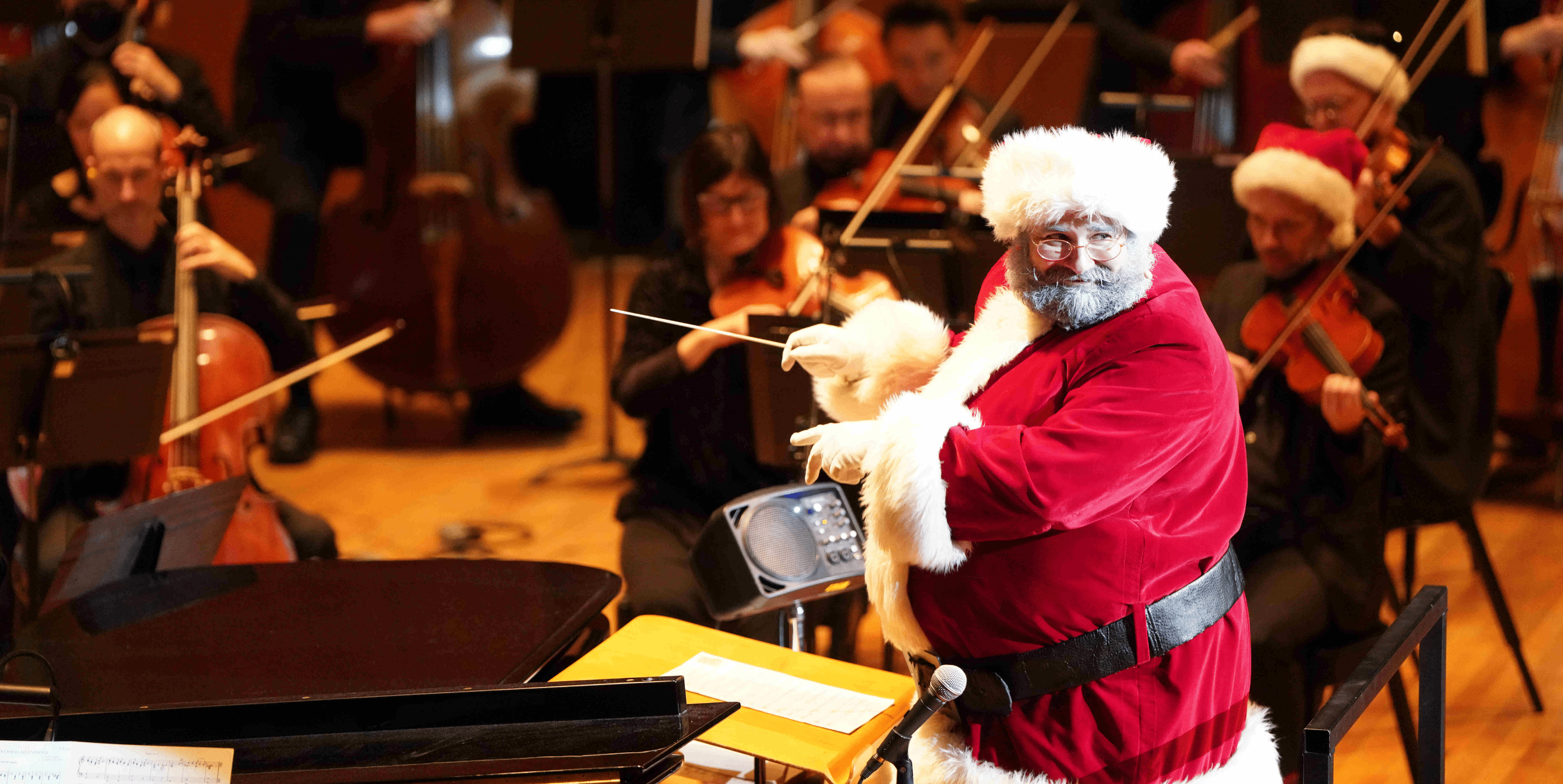 A person dressed as Santa Claus conducts an orchestra, with musicians playing string instruments in the background. The setting is festive and lively, with warm lighting on the stage.