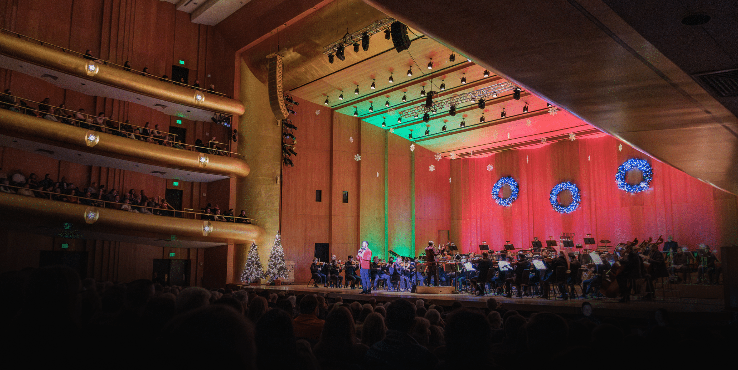 A symphony orchestra performs on a brightly lit stage decorated with festive wreaths and Christmas trees, while an audience watches from the darkened seats of a large concert hall.