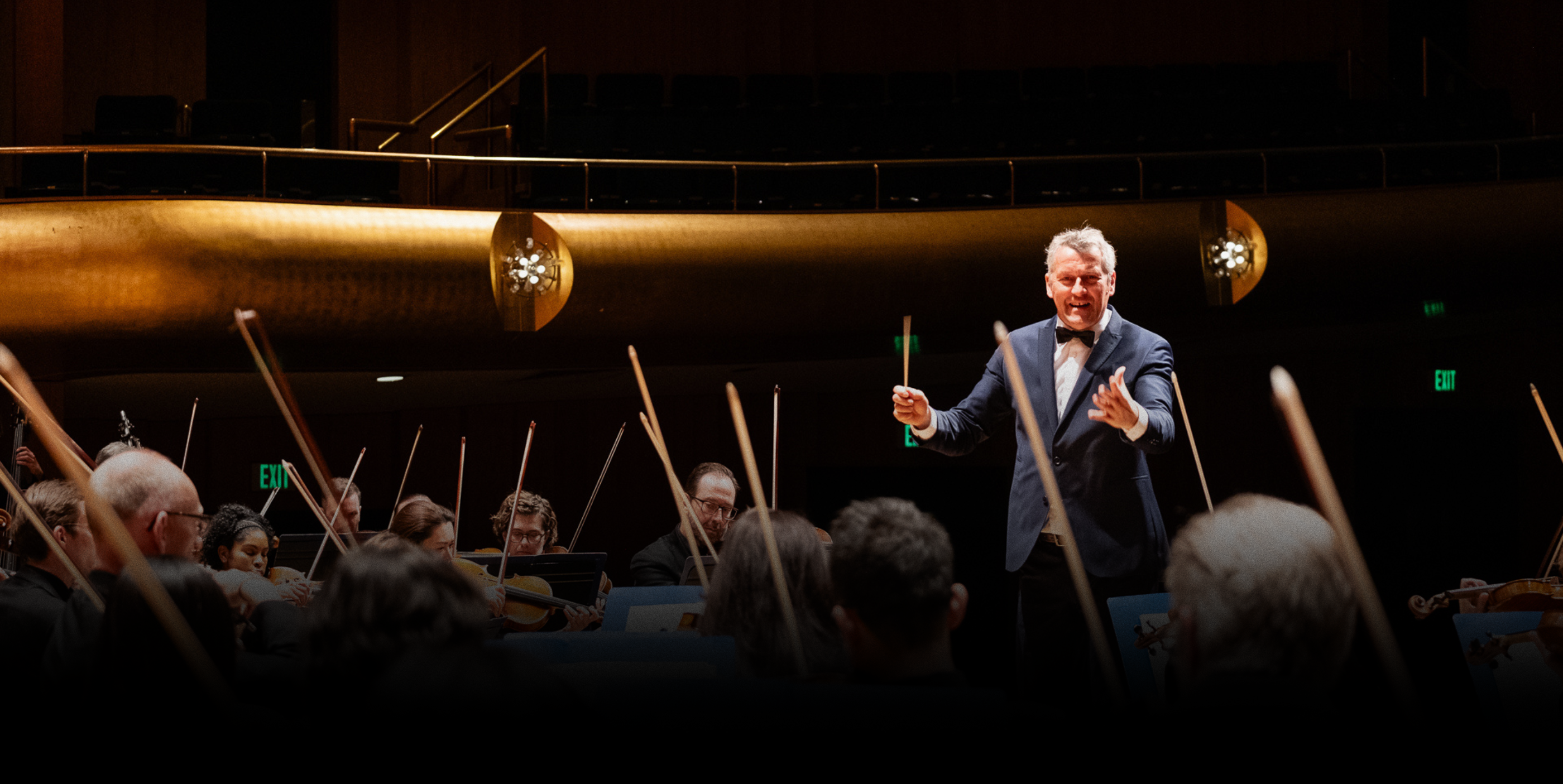 A conductor in a tuxedo energetically leads an orchestra, with musicians playing string instruments in a warmly lit concert hall.