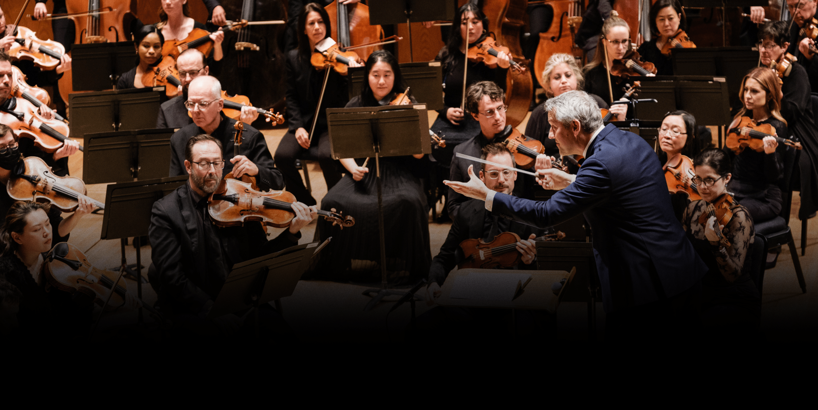 A conductor leads an orchestra of violinists and musicians on stage, all focused on their sheet music and instruments during a classical performance.