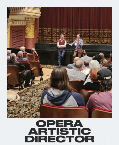 Two men stand on stage speaking to an audience seated in a historic theater with ornate decor and a red curtain. The words Opera Artistic Director are written at the bottom of the image.