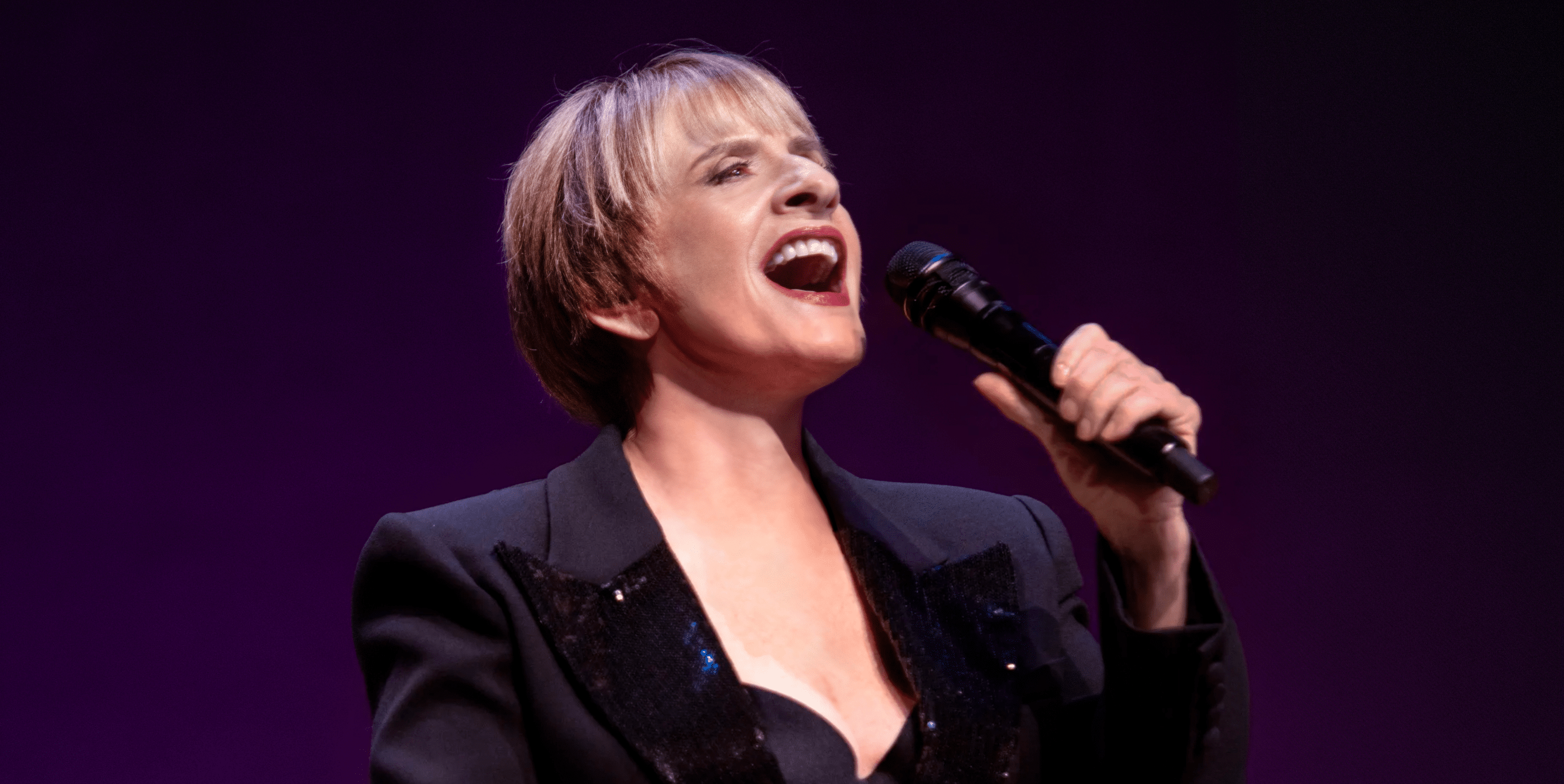 A person with short blond hair sings passionately into a microphone, wearing a dark blazer with a sparkly lapel against a purple background.