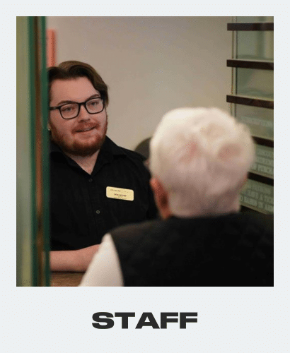A young man with glasses and a name tag smiles while speaking to an older person with short white hair, seen from behind. The word STAFF is displayed at the bottom of the image.