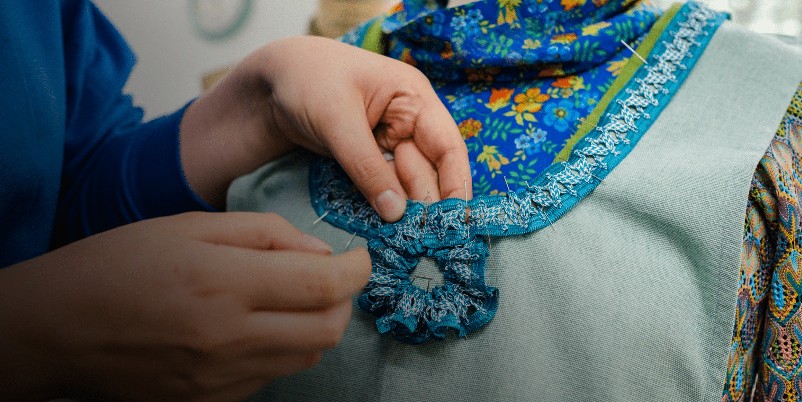 Close-up of hands sewing a decorative blue lace trim onto the neckline of a garment on a dress form, with colorful fabric visible in the background.