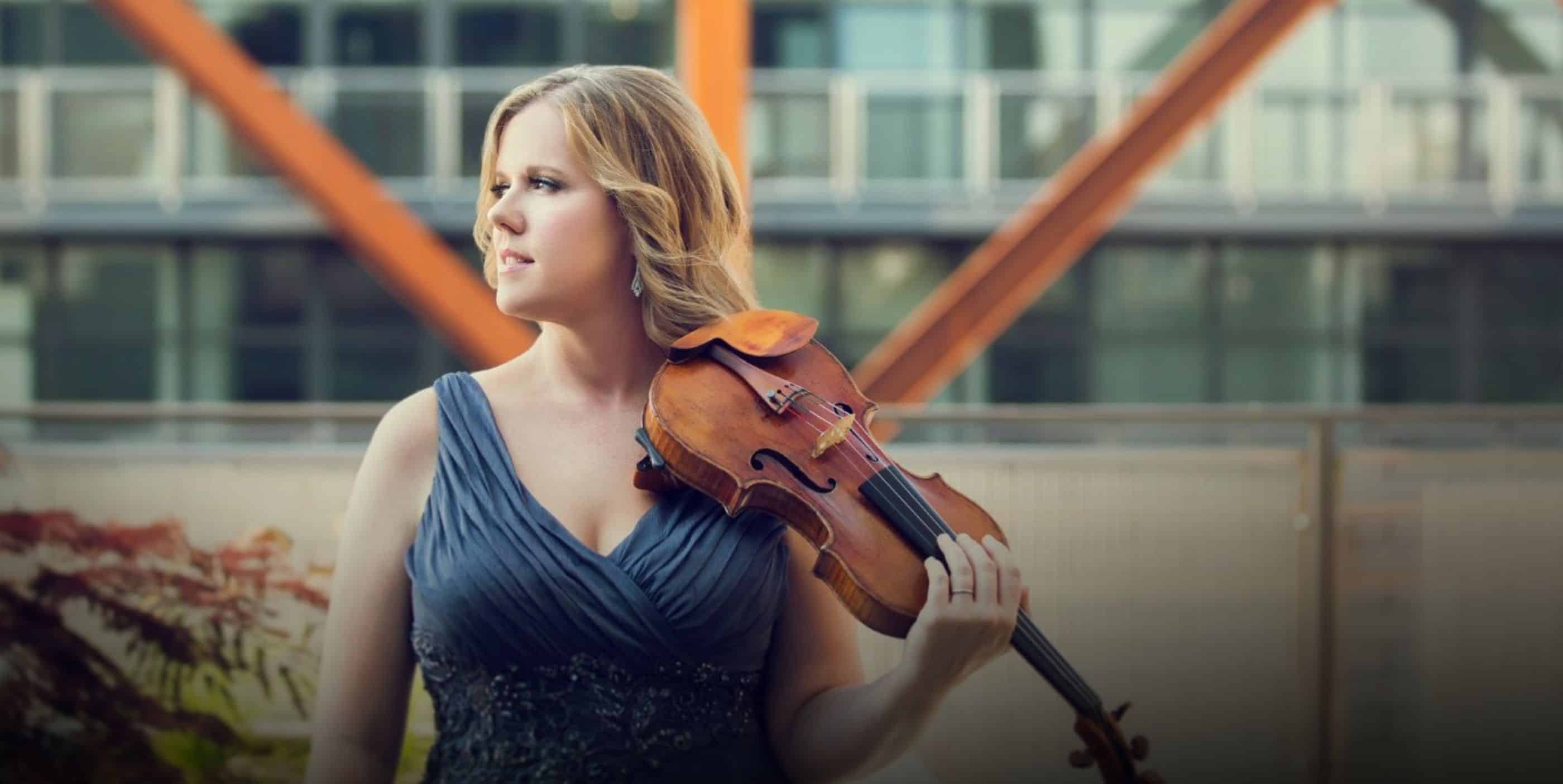 A woman in a sleeveless dark dress stands outdoors, holding a violin on her shoulder and looking to the side, with a modern building and orange beams in the background.