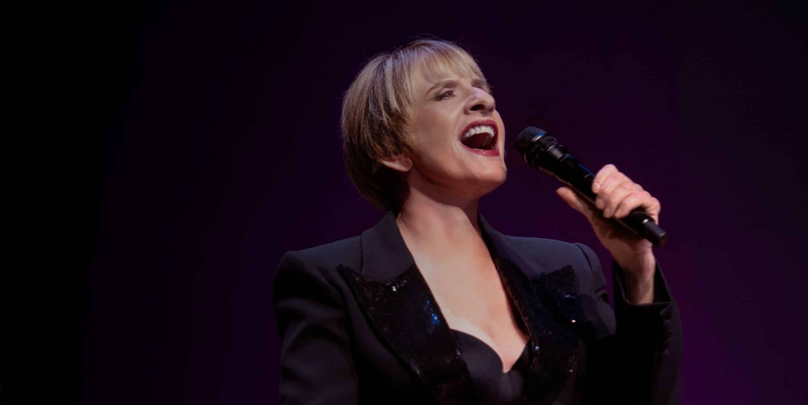A woman with short blonde hair sings passionately into a microphone. She wears a black blazer and stands against a dark, purple-lit background.