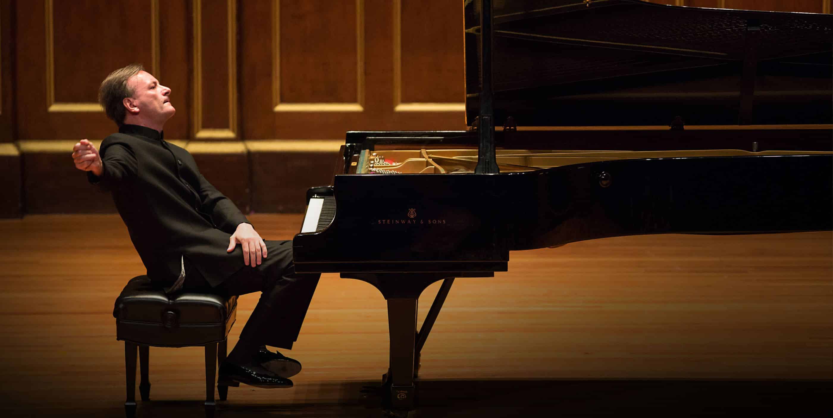 A pianist in formal attire passionately plays a grand piano on stage, leaning back with one arm extended as he performs in a wooden concert hall.