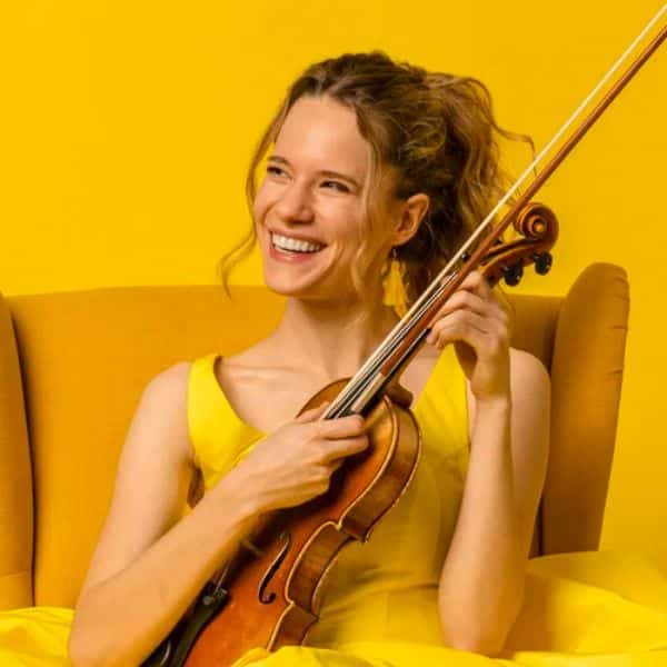 A smiling woman in a yellow dress sits on a yellow chair, holding a violin against a bright yellow background.