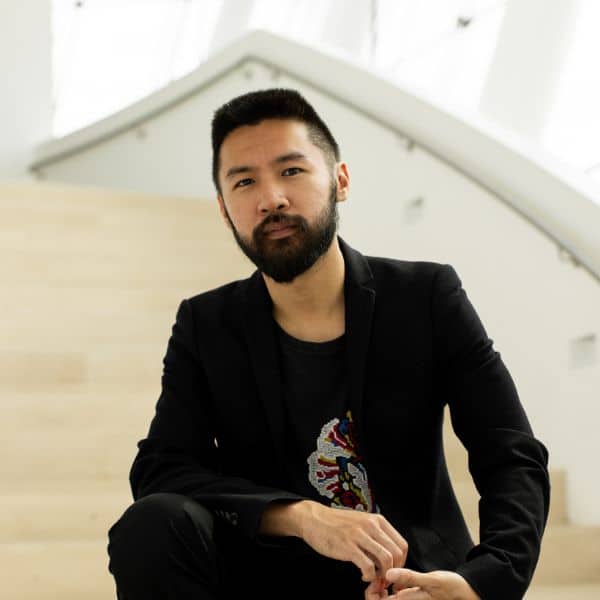 A man with short dark hair and a beard, wearing a black blazer and patterned shirt, sits on light-colored wooden stairs in a bright, modern indoor setting.