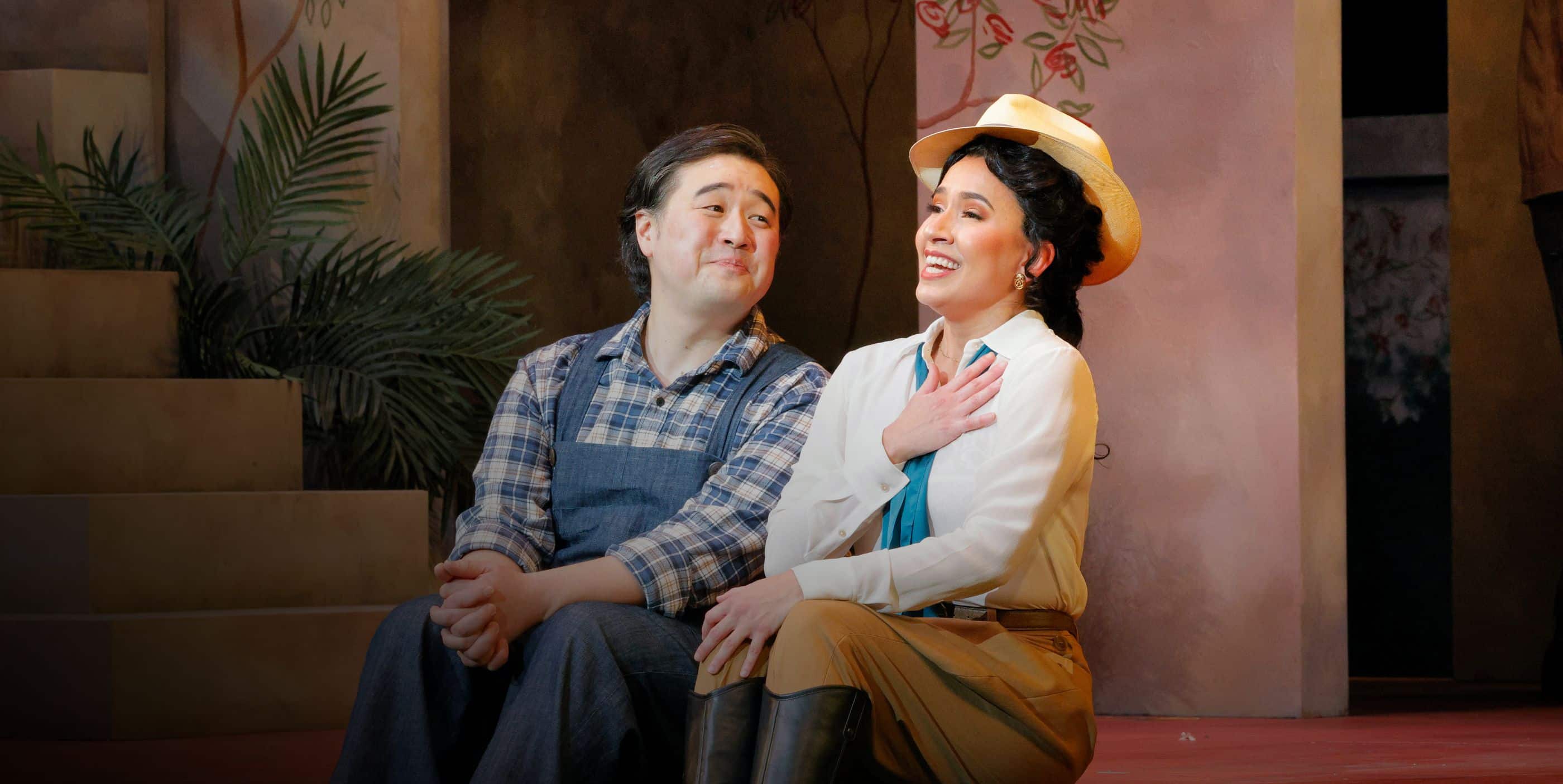 A man in overalls sits on steps next to a smiling woman in a hat and riding clothes, both looking happy under stage lights with plants and painted scenery in the background.
