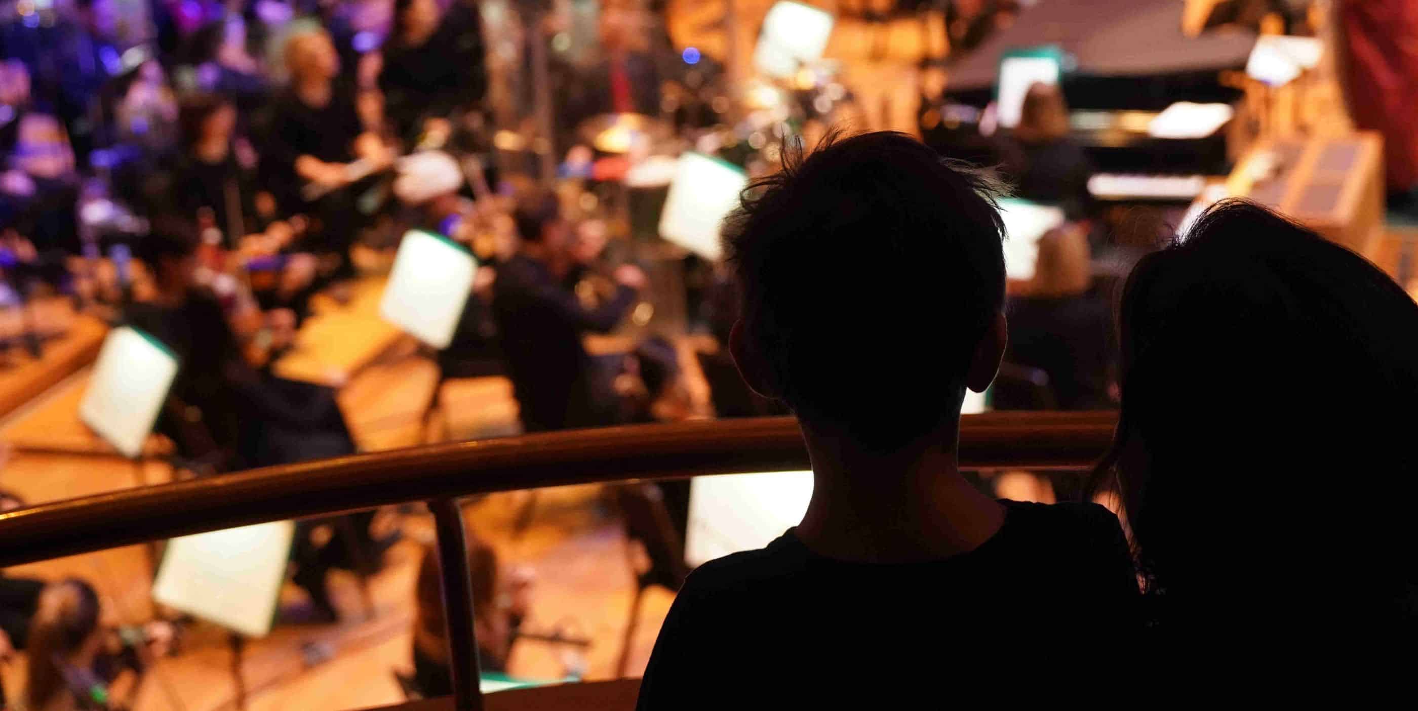 Two people, seen from behind in silhouette, watch an orchestra perform on stage, with musicians and music stands visible in the brightly lit background.