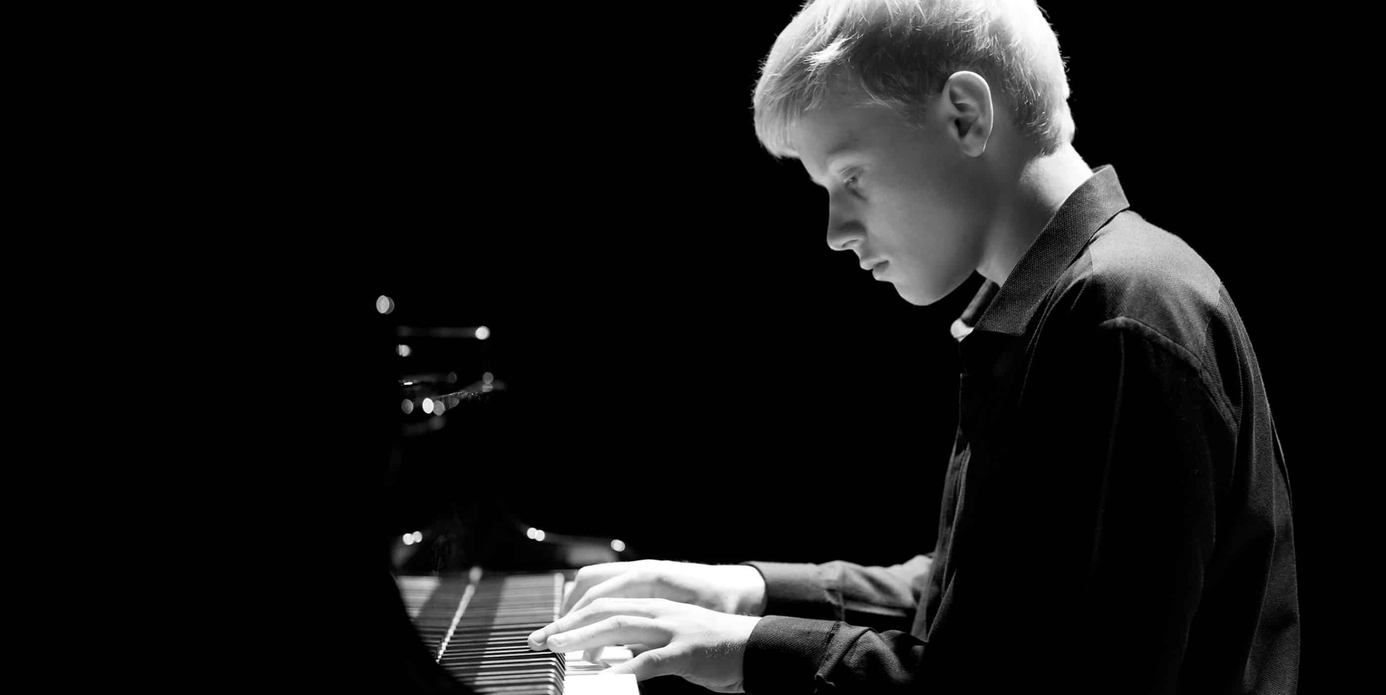 A young person with light hair plays a grand piano, focused intently on the keys. The image is in black and white with dramatic lighting highlighting their face and hands.