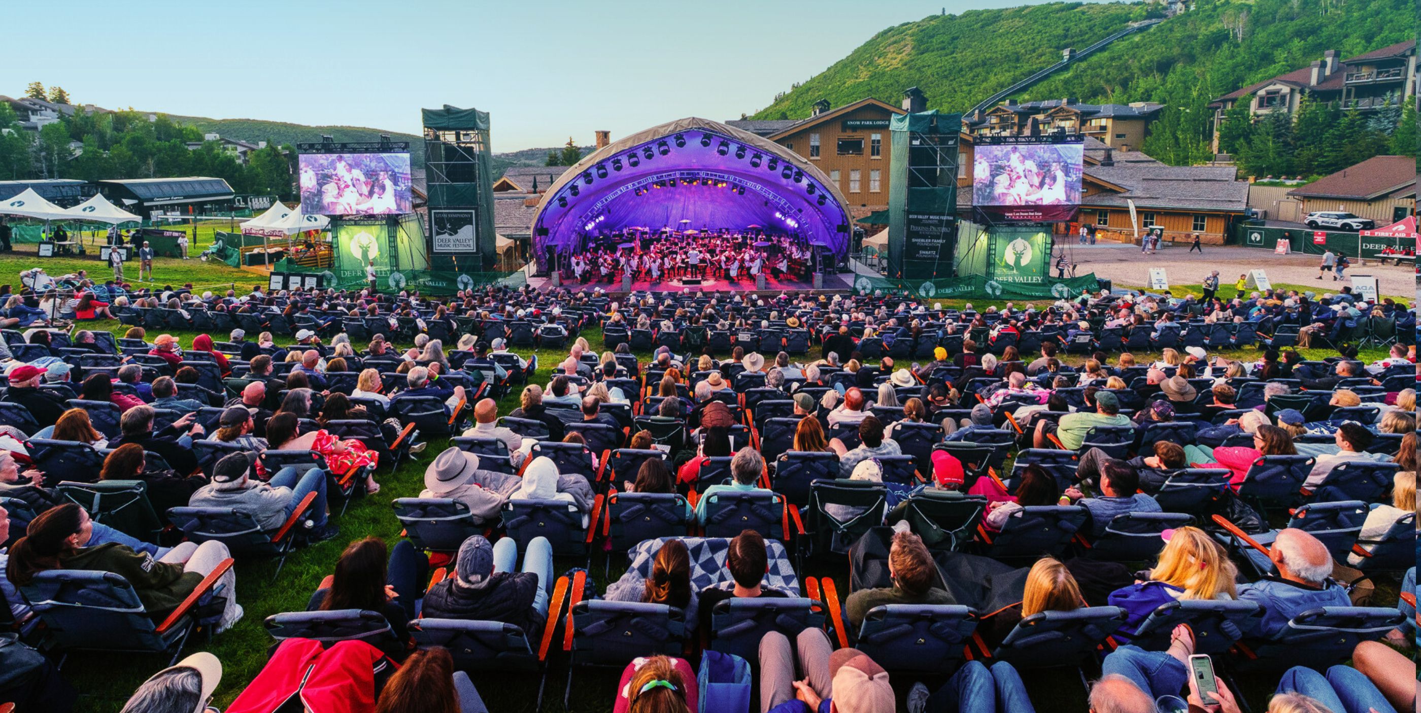 A large outdoor audience sits on lawn chairs facing a stage with purple lighting, where an orchestra performs. Two large screens flank the stage, and mountains and buildings are visible in the background.