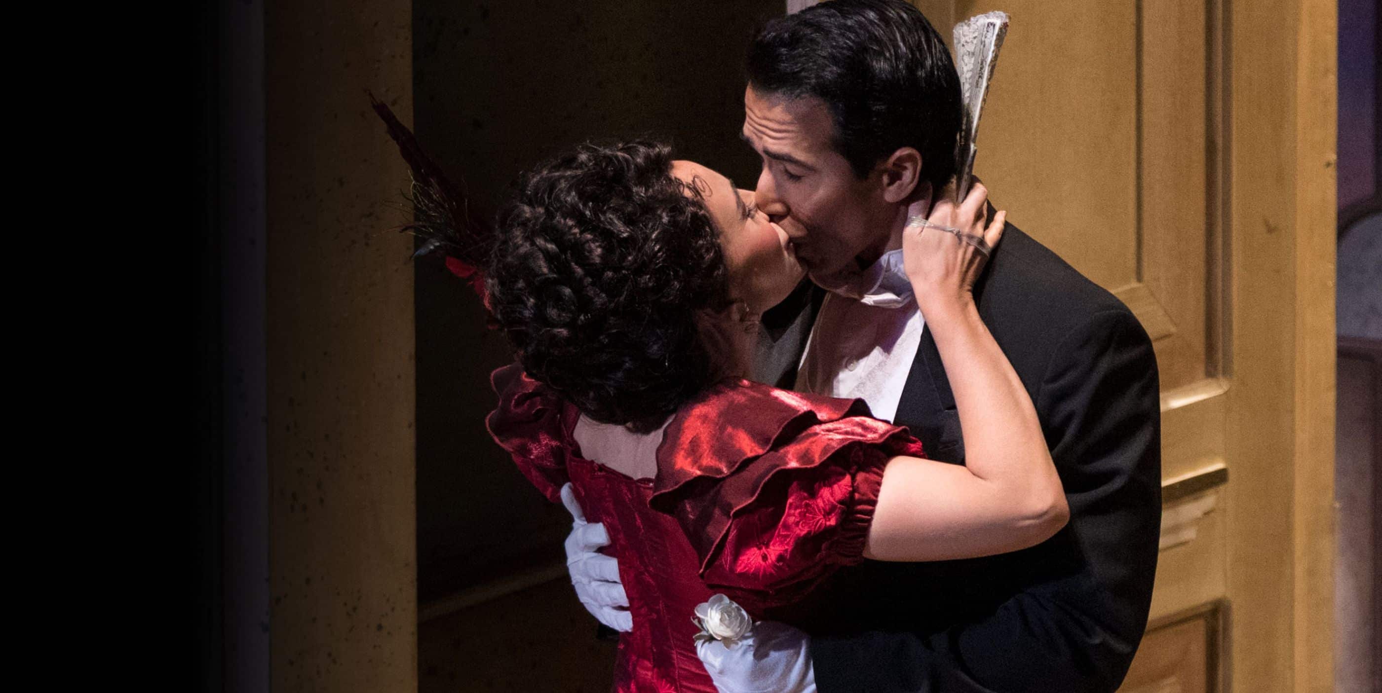 Two people in formal attire share a passionate kiss; the woman wears a red dress with gloves and holds a fan, while the man is in a black suit and white bow tie. They embrace against a backdrop of wooden panels.