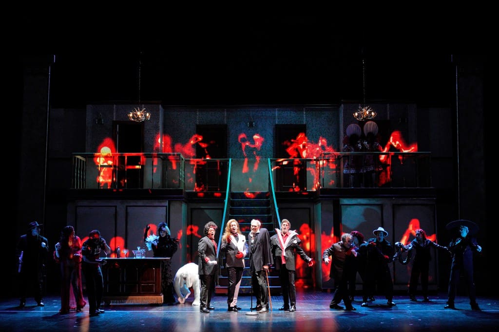 Dozens of people stand on a stage with a two-level set piece and a large staircase. Red lighting obscures most of the people on stage, but four men in black suits stand at the front of the stage, singing under a spotlight.