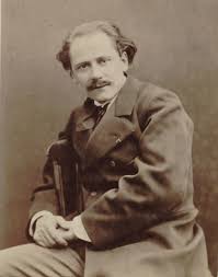 Middle-aged man, receding hairline, moustached, looking at the camera while seated sideways with arm over the back of a chair.