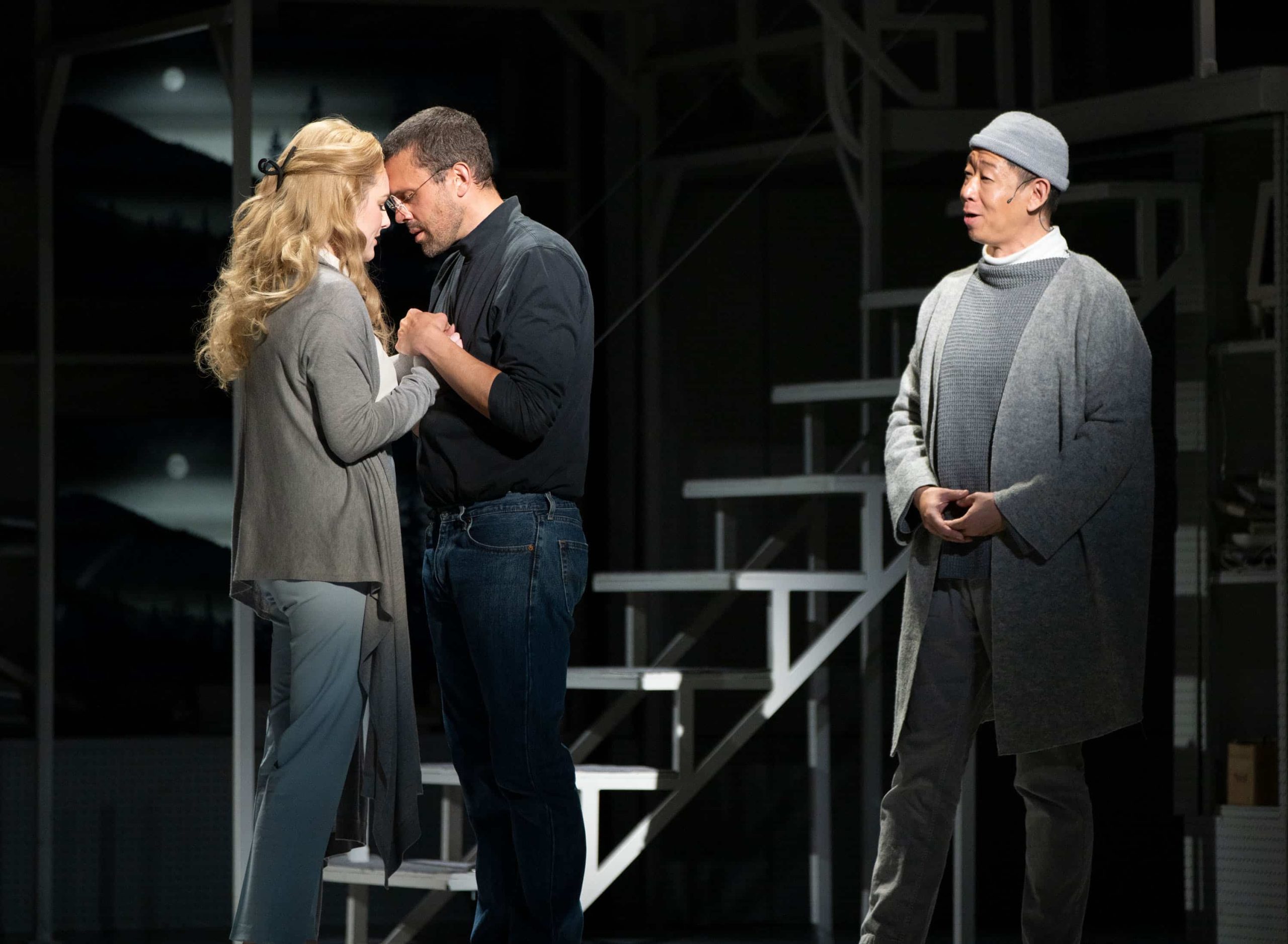 A woman and a man face each other holding hands with their foreheads touching. The woman has blond hair; the man is wearing a black turtleneck. Behind them, a man wearing all grey watches them with his mouth open as if singing.