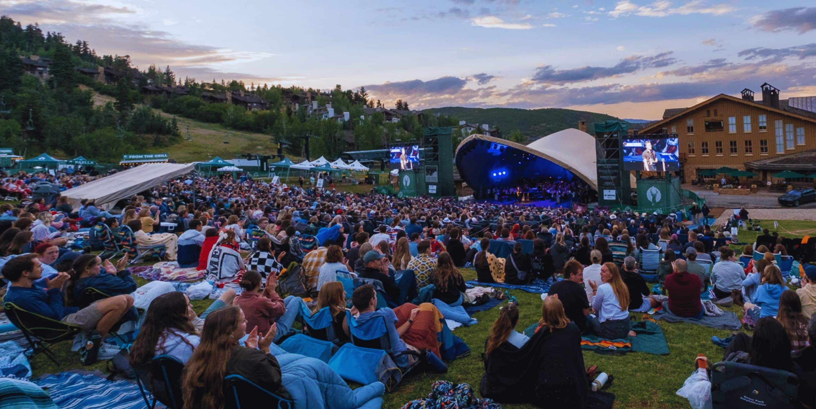 A large crowd sits on blankets and chairs on a grassy hillside, watching an outdoor concert on a stage with blue lights. Trees, tents, and a lodge are visible in the background under a partly cloudy sky at sunset.