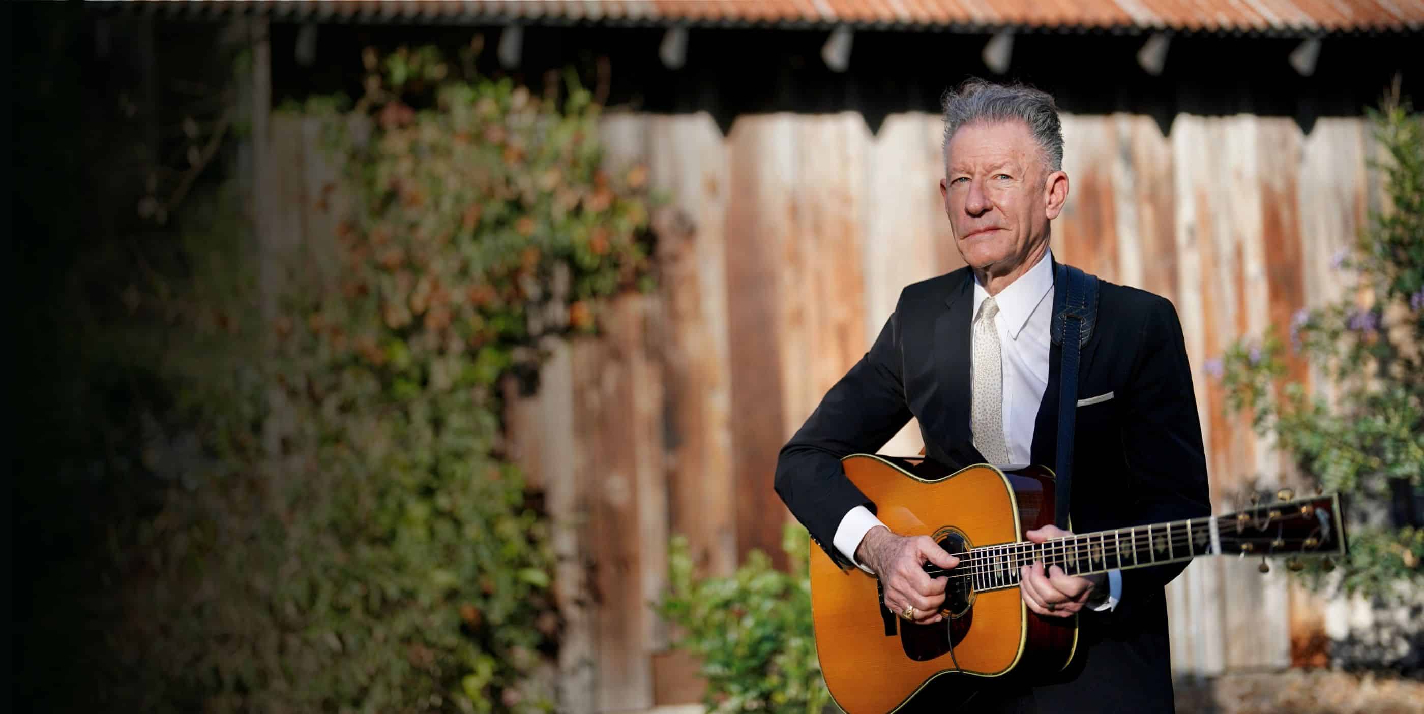 A man in a black suit and white tie stands outdoors, holding an acoustic guitar. He is positioned in front of a weathered wooden building with greenery visible around him.