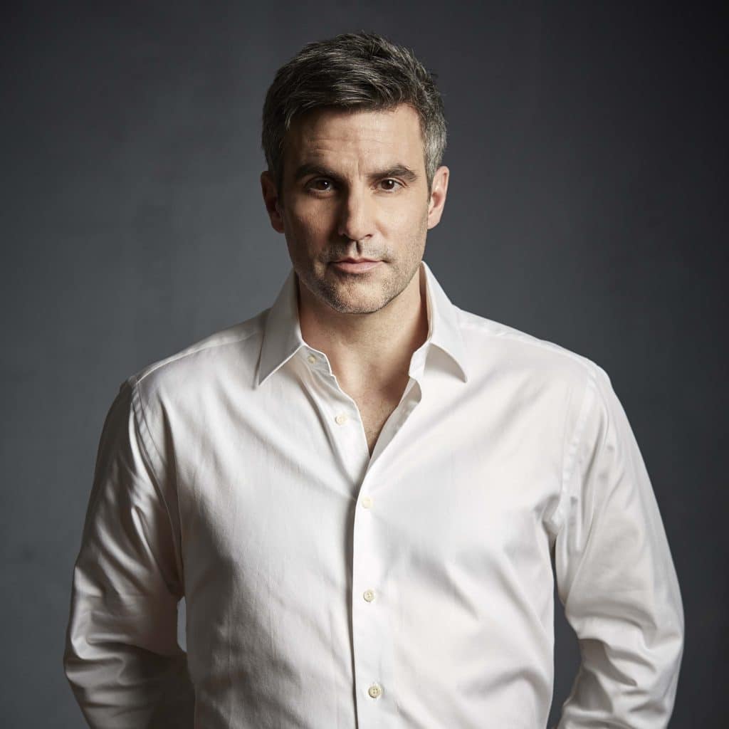 A man with short gray hair wearing a white button-up shirt stands against a dark gray background, looking directly at the camera with a neutral expression.