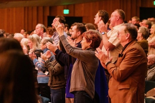 A group of people stand and clap in an auditorium, giving a standing ovation. The audience appears engaged and enthusiastic, with warm lighting highlighting their faces and expressions.
