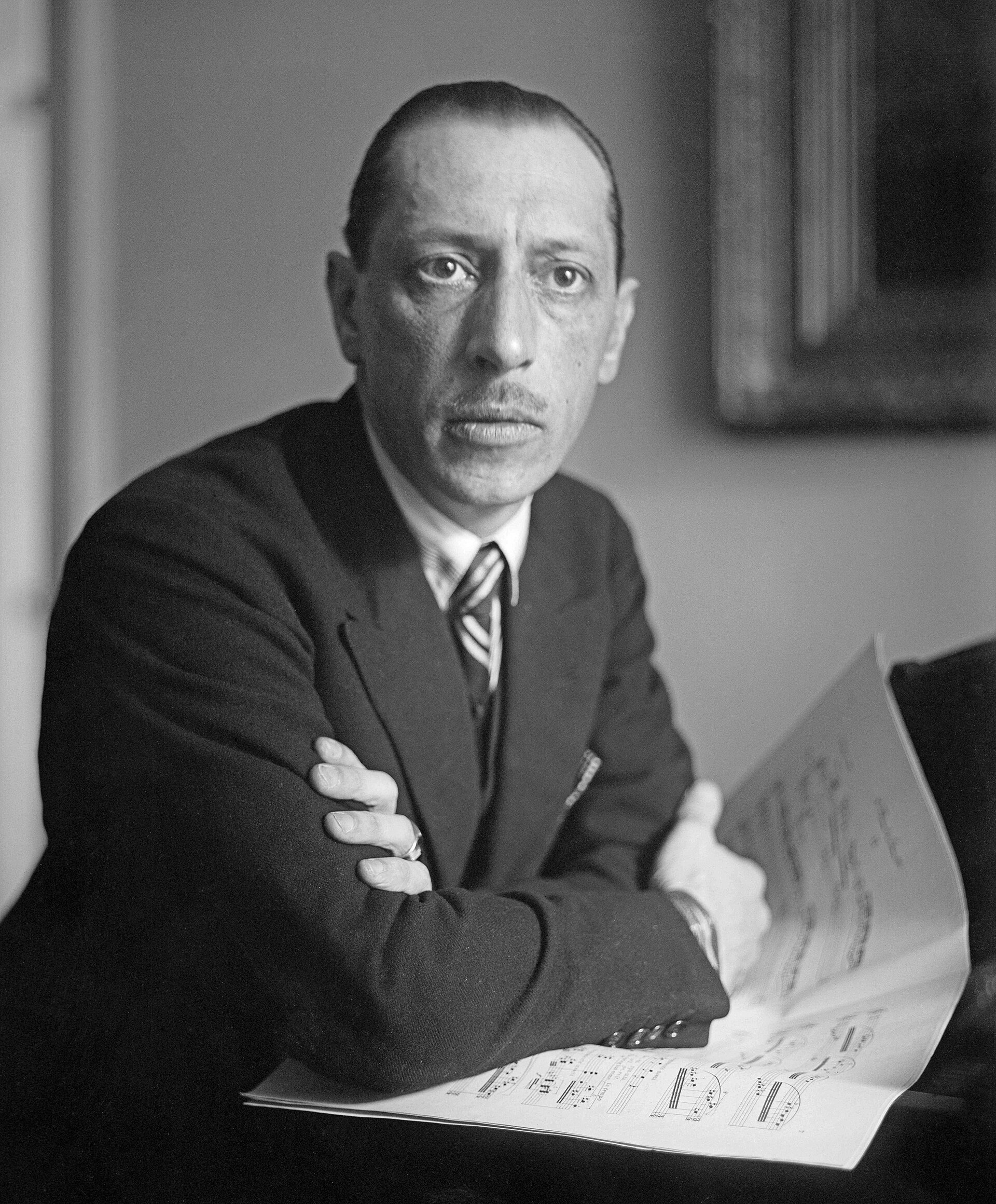 A man in a suit with a tie sits holding sheet music, looking directly at the camera. He has neatly combed hair and a serious expression, with his arms crossed. The background is softly lit and unfocused.