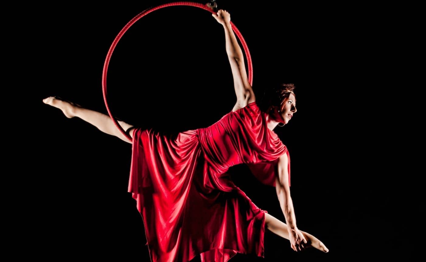 A woman in a flowing red dress performs an aerial split on a suspended hoop against a black background, showcasing strength and grace.
