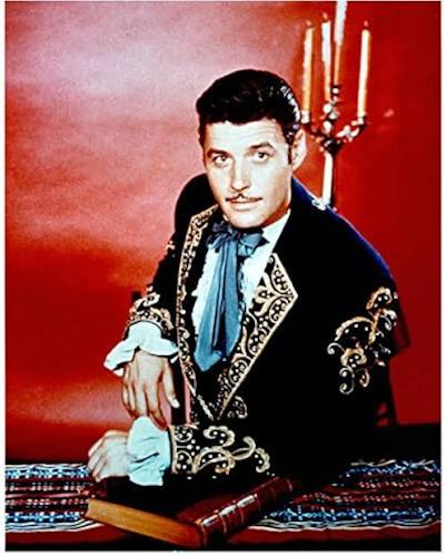 A man in a dark jacket with gold embroidery and light blue necktie leans forward on a large book. Behind him is a red backdrop with a candelabra holding lit candles.