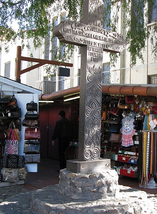 An intricately carved cross serves as a monument to the founding of Los Angeles. The cross has the inscription, "El Pueblo de Nuestra Señora la reina deLos Ángeles. Felipe de Neve. September Fourth 1781." Behind the monument are vendor stalls selling various items.