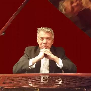 A man with grey hair sits behind a polished brown piano, resting his chin on his clasped hands. He is wearing a dark suit jacket over a white shirt and is looking directly at the camera. In the upper right corner, a soft reflection of his face is visible on the underside of the piano's raised lid. The background behind him is a solid dark red.
