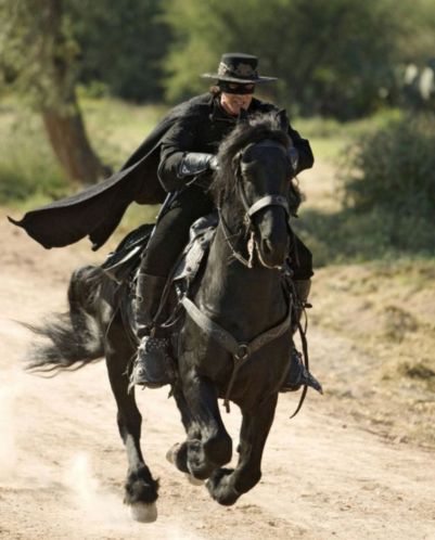A mansked rider in black gallops on a dark horse along a dusty path.