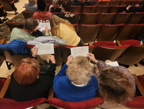 Audience members in a theater hold printed programs and read them.