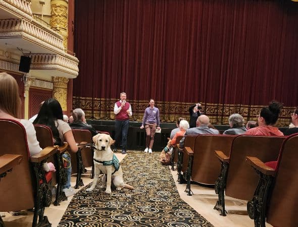 A theater audience faces two people speaking on stage. A guide dog sits in the aisle, wearing a harness.