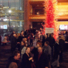 A group of people socializing in a large, vibrant lobby with a tall, intricate red glass sculpture as a centerpiece. The space is well-lit, with modern architecture and large windows in the background.